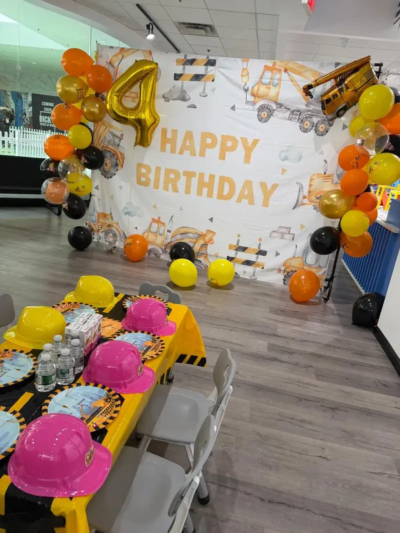 The party room set for a construction birthday, with a Happy Birthday backdrop, a balloon arch and hard hats on the table